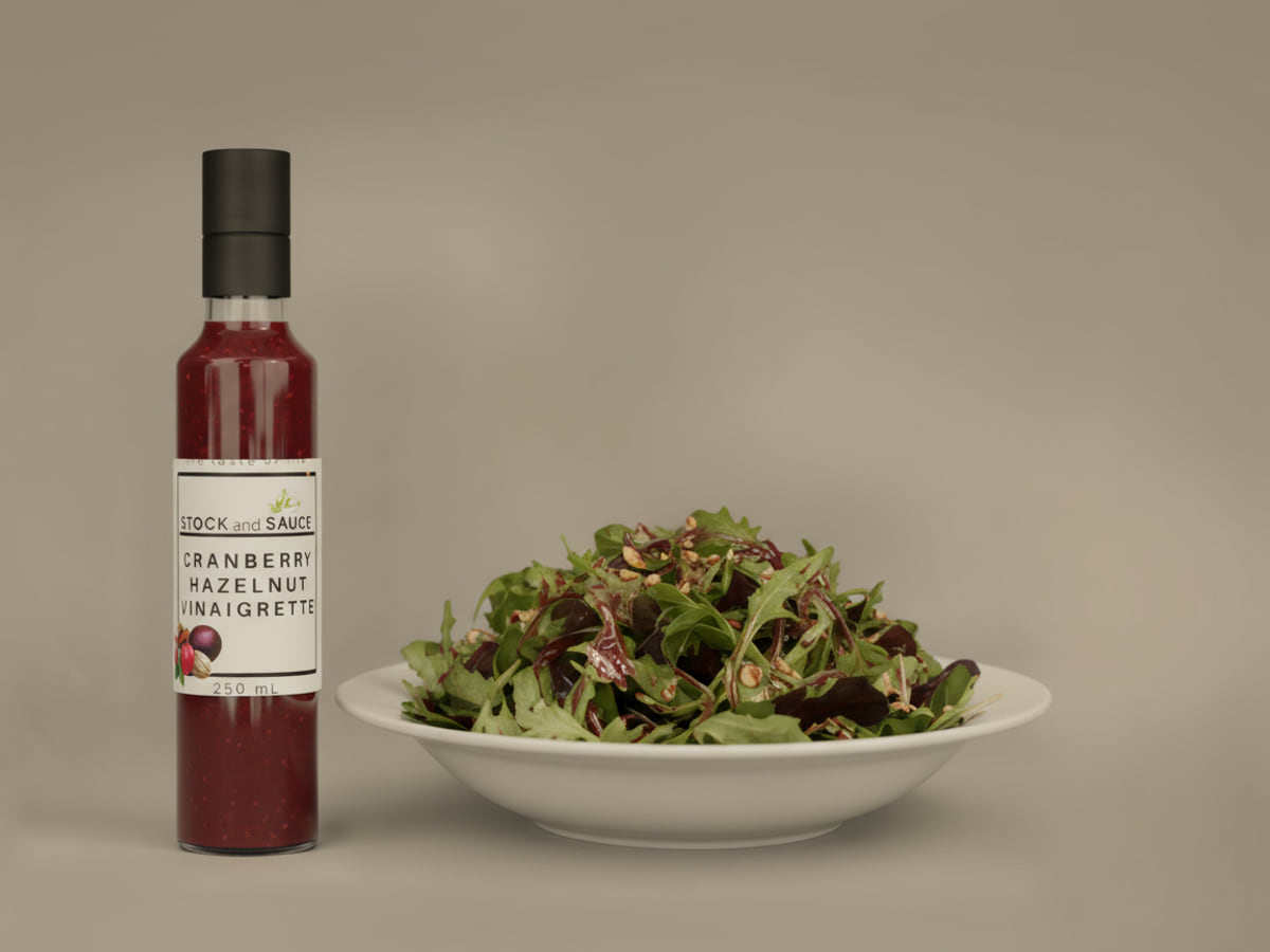 Bottle of Stock & Sauce Cranberry Hazelnut Vinaigrette, a deep red-brown dressing chunky with pale nut pieces, beside a bowl of mixed leaves dressed with it