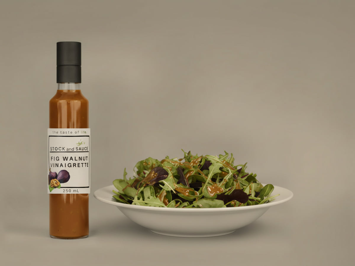 Bottle of Stock & Sauce Fig Walnut Vinaigrette, a dark amber-brown dressing flecked with pale seeds, beside a bowl of mixed leaves dressed with it