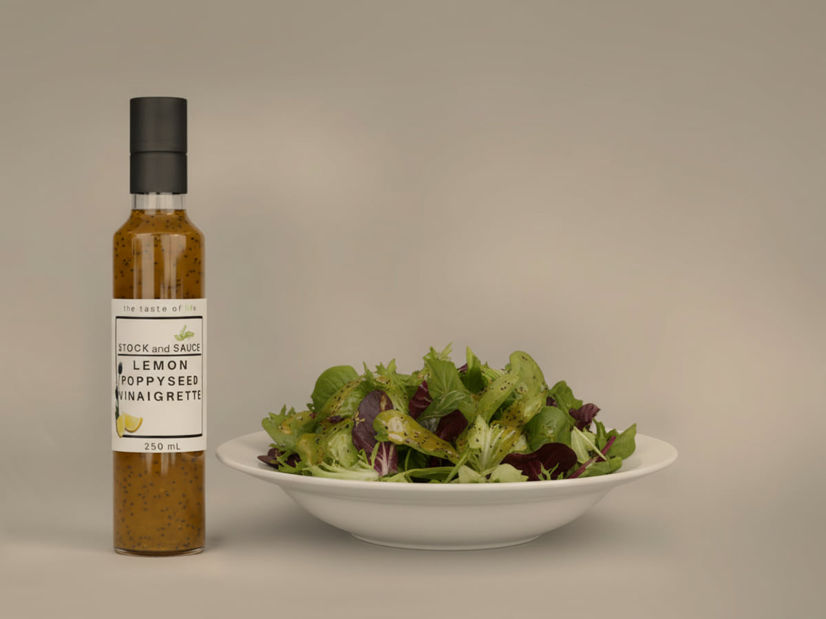 Bottle of Stock & Sauce Lemon Poppyseed Vinaigrette, golden and flecked with poppyseed, beside a bowl of mixed leaves dressed with it