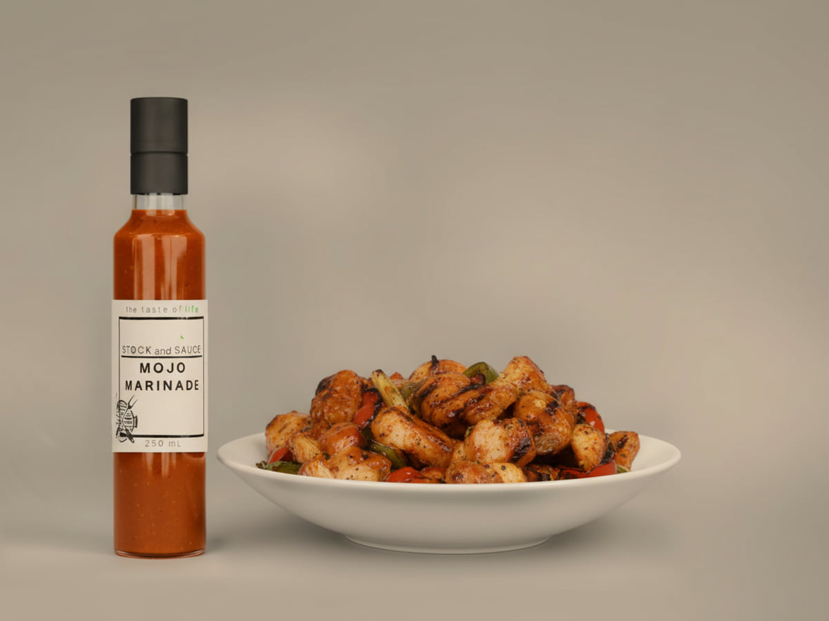 Bottle of Stock & Sauce Mojo Marinade, a thick deep rust-orange citrus-garlic marinade flecked with garlic and pepper, beside a bowl of grilled meat and peppers glazed with it